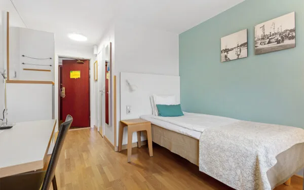 Enkelrum med enkel säng, ljusgrön fondvägg, skrivbord och trägolv.
