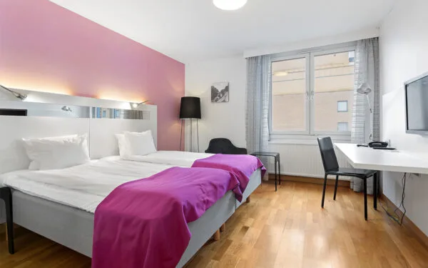 Hotellrum med två enkelsängar, rosa fondvägg, skrivbord med stol och stort fönster.