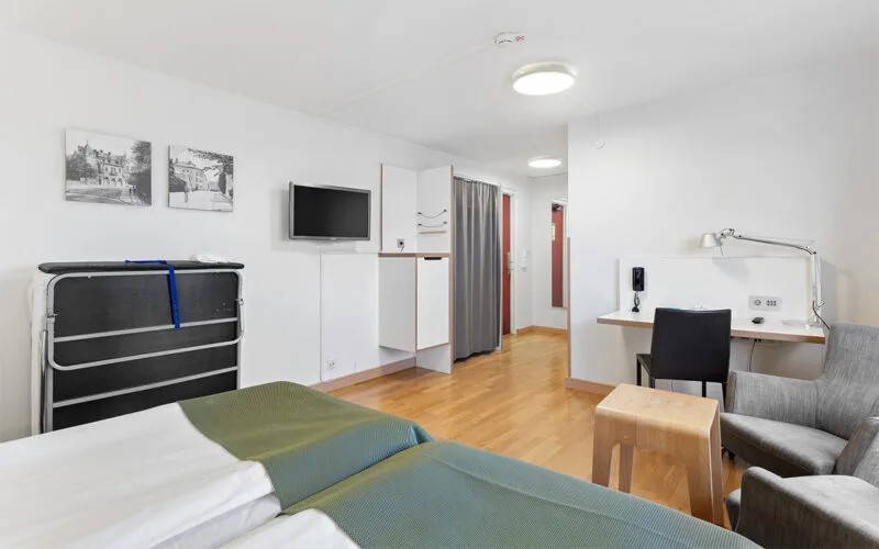 Hotellrum med dubbelsäng, skrivbord, TV, sittgrupp och extrasäng mot väggen.