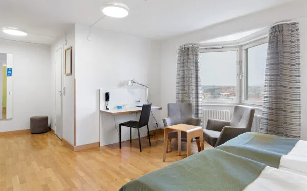 Hotellrum med två sängar, skrivbord, sittgrupp och stort fönster med utsikt över staden.