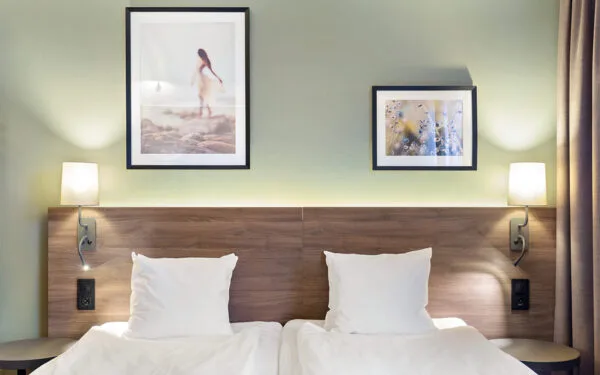 Hotellsäng med två kuddar, träfärgad sänggavel, två vägglampor och tavlor ovanför.