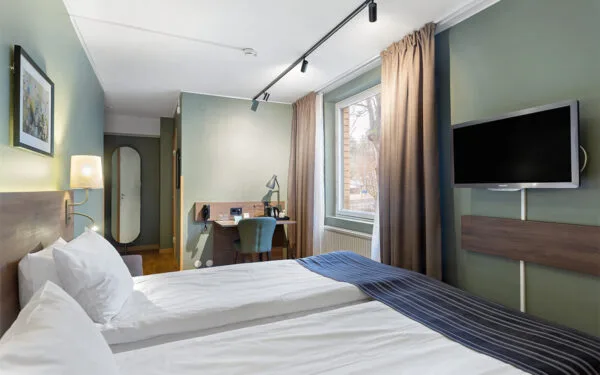 Hotellrum med dubbelsäng, skrivbord, fåtölj, vägg-TV och stort fönster med gardiner.