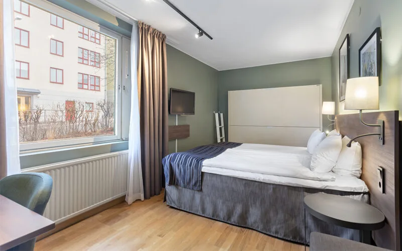 Hotellrum med dubbelsäng, grön vägg, skrivbord och stort fönster med utsikt.