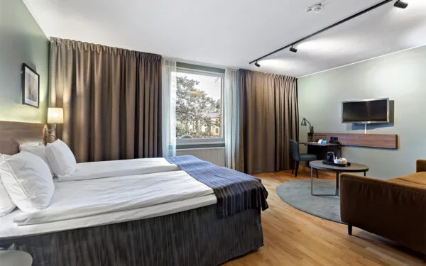 Hotellrum med dubbelsäng, skrivbord, TV och sittgrupp vid ett stort fönster.