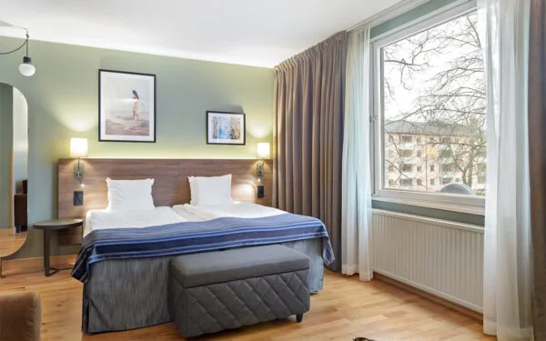 Hotellrum med dubbelsäng, grön fondvägg, nattduksbord och stort fönster med gardiner.