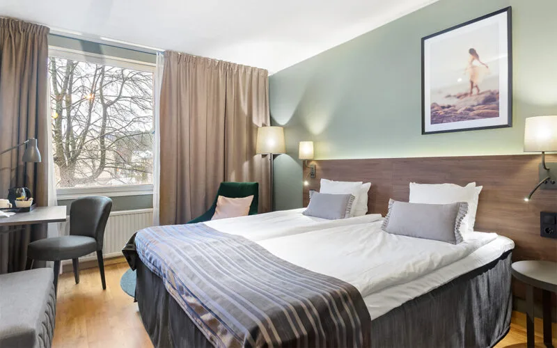 Hotellrum med två bäddar, skrivbord, fåtölj vid fönstret och tavla ovanför sängen.