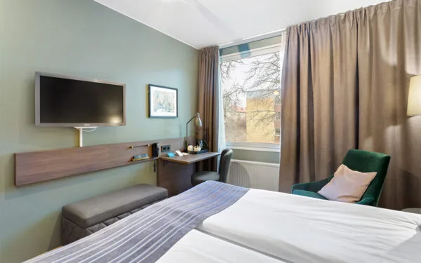 Hotellrum med säng, skrivbord med arbetsyta, TV på väggen och fåtölj vid fönstret.