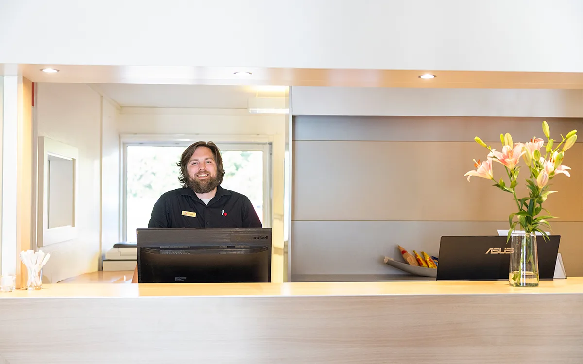 Leende receptionist bakom hotellreception med blommor på disken.