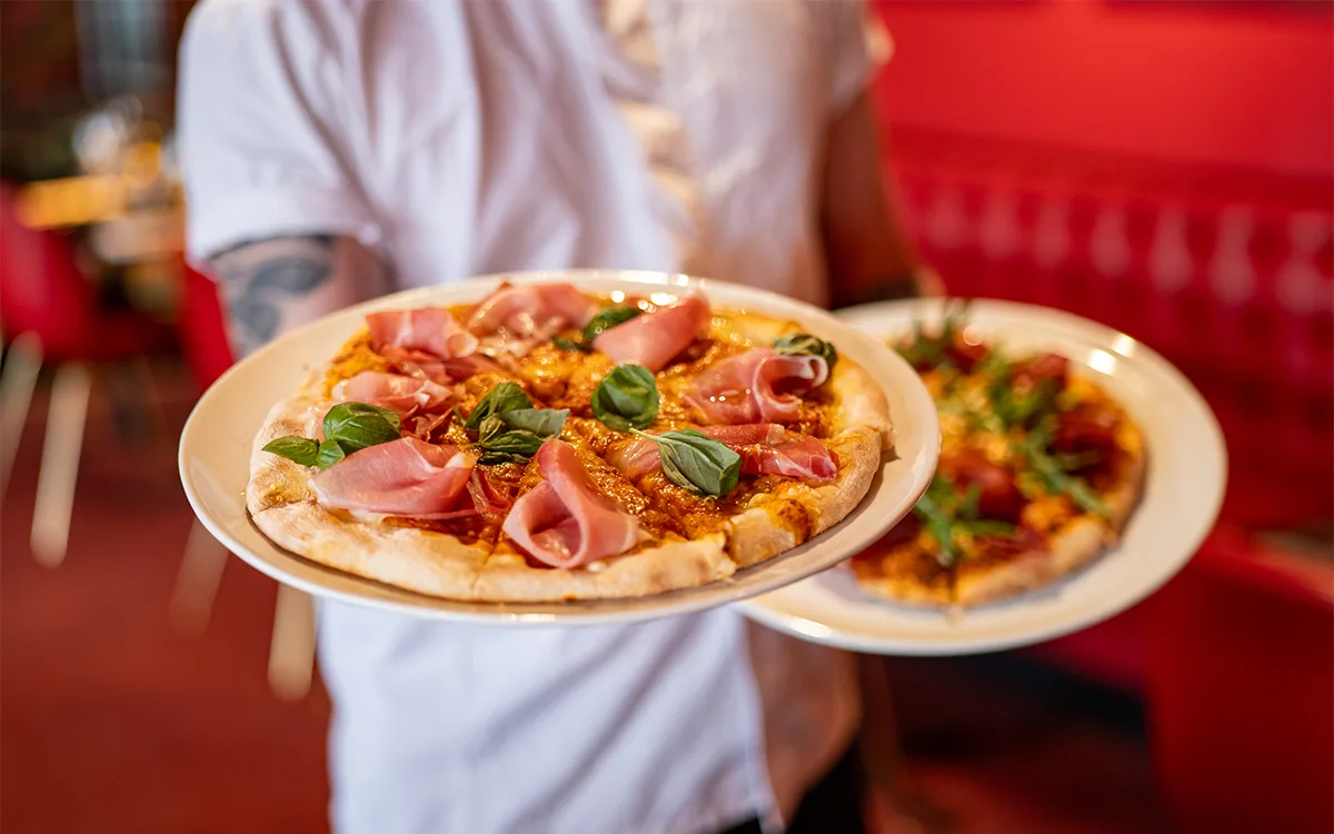 Servitör håller tallrikar med nygräddade pizzor toppade med skinka och basilika.