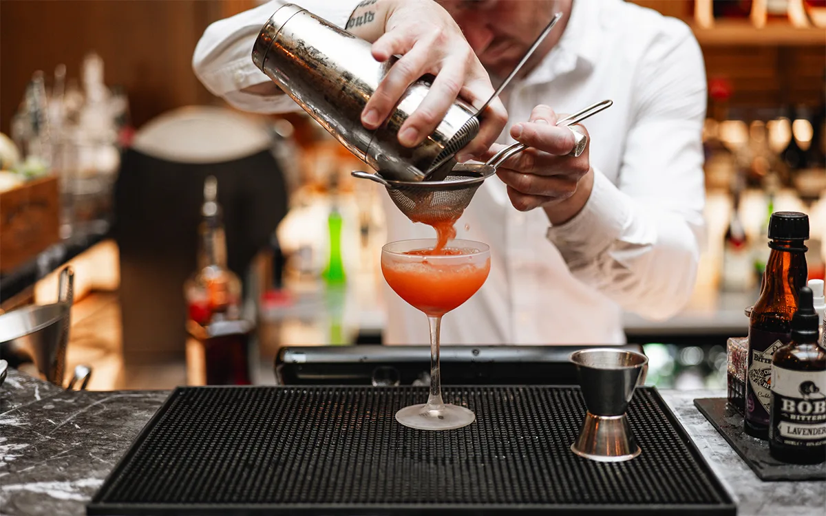 Bartender häller upp en röd cocktail i glas genom sil.