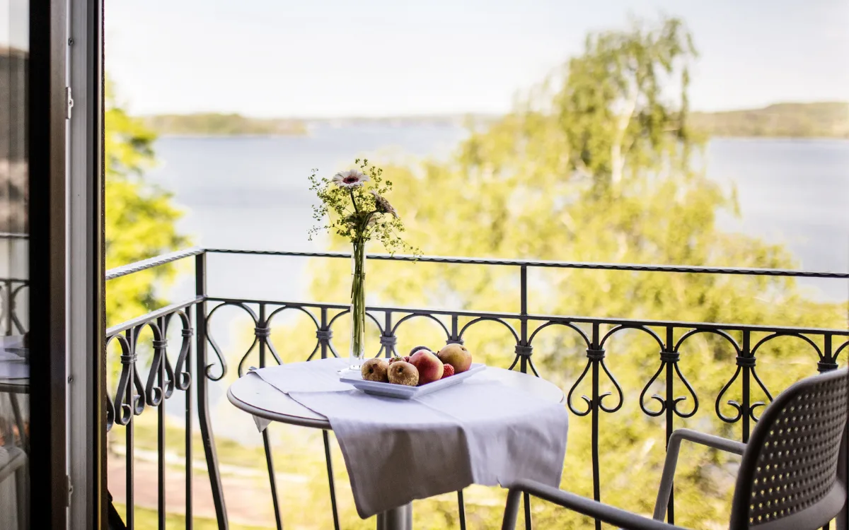 Balkon med bord med frugt og kager, udsigt over sø og grønne områder.