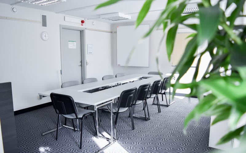 Konferensrum med långbord, svarta stolar och whiteboard i ljus miljö med växt i förgrunden.