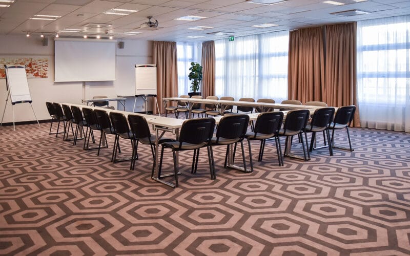 Konferensrum med U-formad möblering, projektorduk och stora fönster med gardiner.