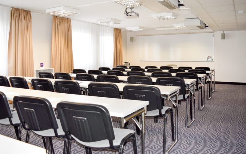 Konferensrum med skolbänksmöblering, projektorduk och beige gardiner.