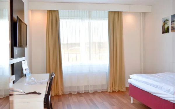 Ljust rum med enkelsäng, arbetsyta och TV på väggen.