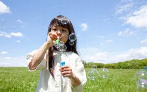 Flicka blåser såpbubblor på en grön äng under blå himmel.