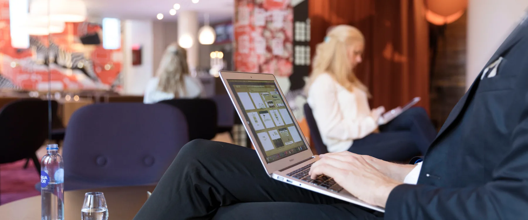 Personer sitter i en hotellobby och arbetar, en med laptop och en annan med anteckningar.