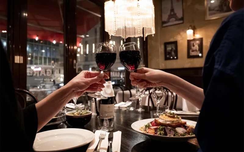 Två personer skålar med glas rött vin vid ett dukat restaurangbord med mat, i en stämningsfull miljö med levande ljus och utsikt över staden