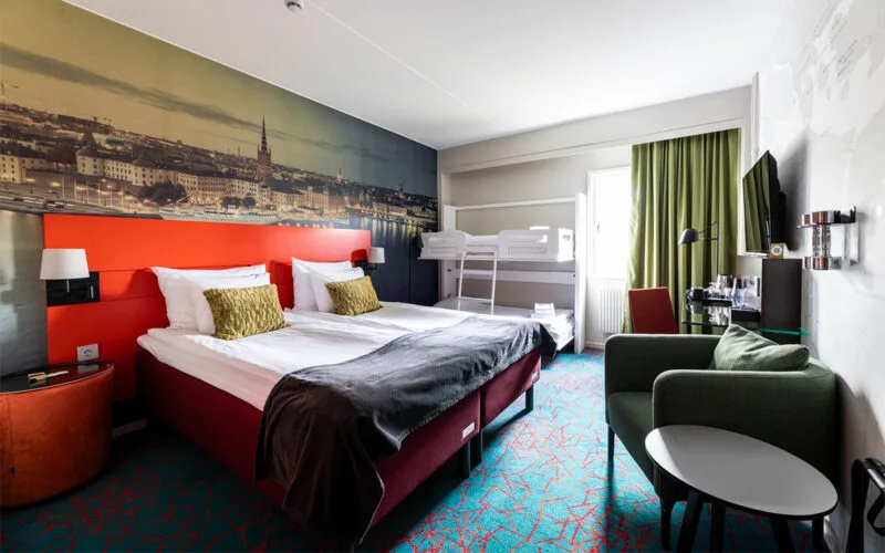 Hotellrum med dubbelsäng, våningssäng och fondvägg med stadsmotiv