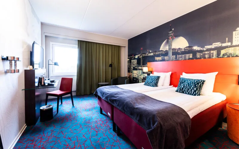 Hotellrum med två enkelsängar bredvid varandra, skrivbord och stol, helteckningsmatta, hantlar och gardiner