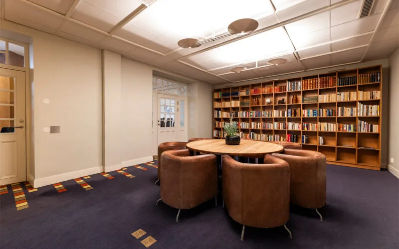 Mötesrum med runda bord, bruna skinnfåtöljer och en stor bokhylla fylld med böcker längs väggen.