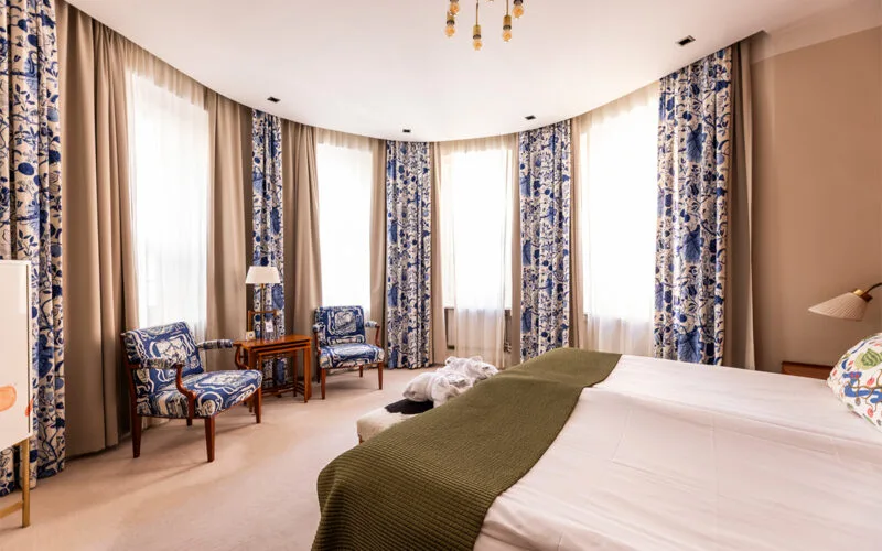 Ljust hotellrum med dubbelsäng, grön filt och blåmönstrade gardiner vid stora fönster