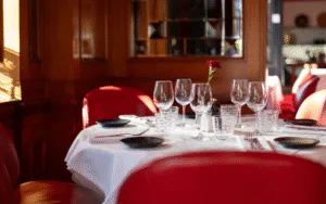 A round table with white tablecloth, glasses and dishes, surrounded by red chairs in an elegant restaurant setting.