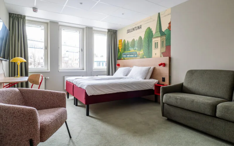 Översikt av tvåbäddsrum med säng, skrivbord, stol, fotölj och bäddsoffa samt heltäckningsmatta.