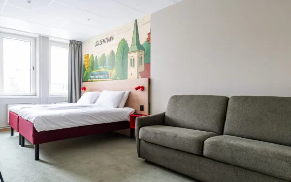 Översikt av tvåbäddsrum med säng, skrivbord, stol, fotölj och bäddsoffa samt heltäckningsmatta.