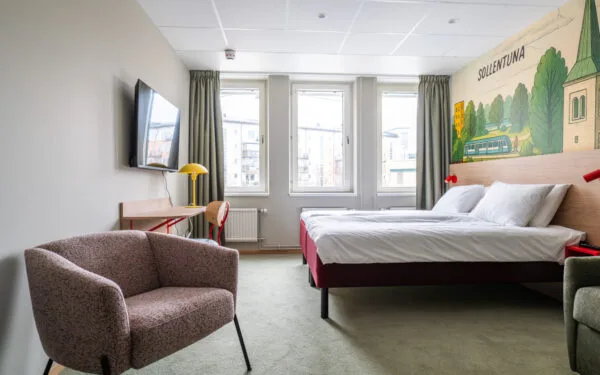 Översikt av tvåbäddsrum med säng, skrivbord, stol, fotölj och bäddsoffa samt heltäckningsmatta.