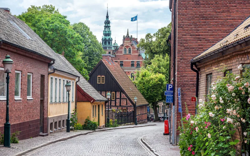 En pittoresk kullerstensgata i Lund med färgglada hus och ett torn från universitetets huvudbyggnad i bakgrunden.