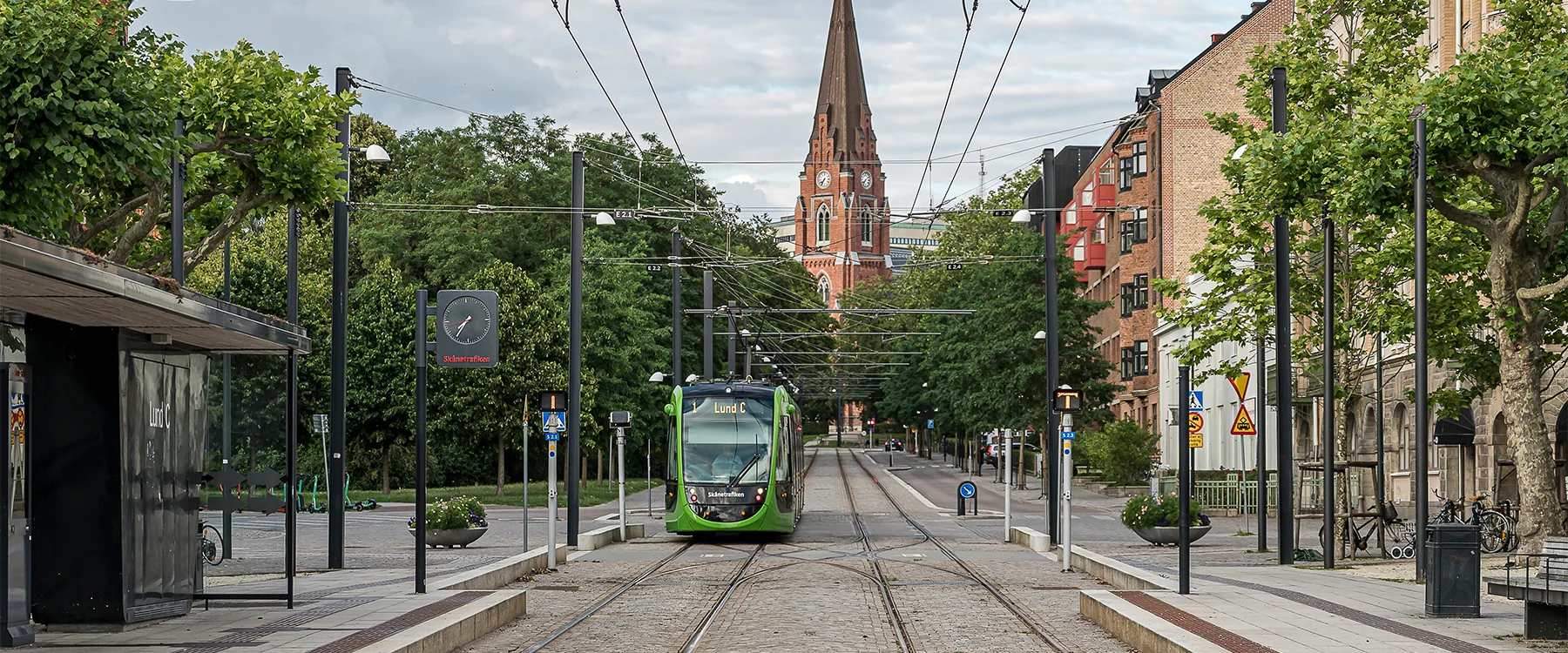 En grön spårvagn kör på spåren i Lund med en kyrka i bakgrunden.