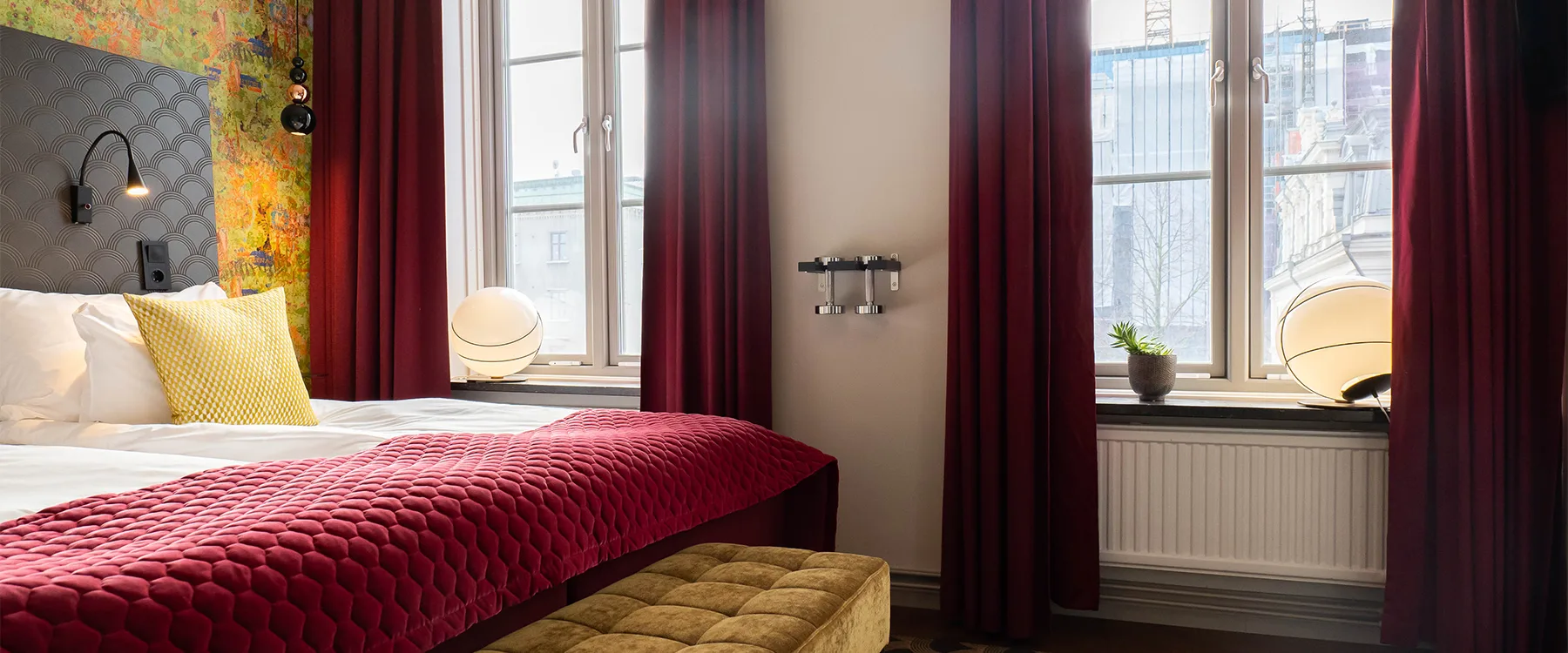Hotellrum med röd bäddad säng, vinröda gardiner och en sittbänk vid fotänden.