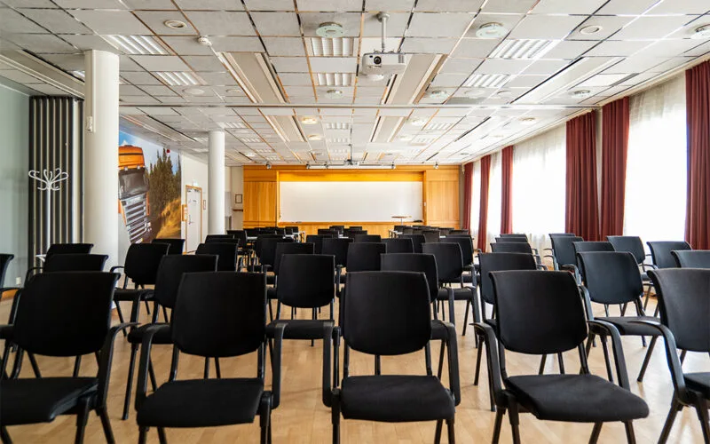 Konferensrum med uppställda bord och stolar, projektor i taket och stor duk framtill.