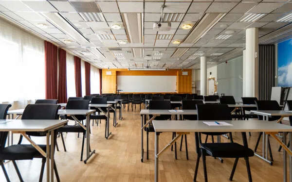 Konferensrum med uppställda bord och stolar, projektor i taket och stor duk framtill.