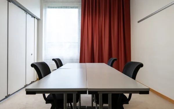 Mötesrum med två bord, svarta stolar, röd gardin och stort fönster.