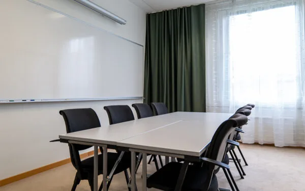 Mötesrum med vitt bord, svarta stolar, whiteboard och stort fönster med gröna gardiner.
