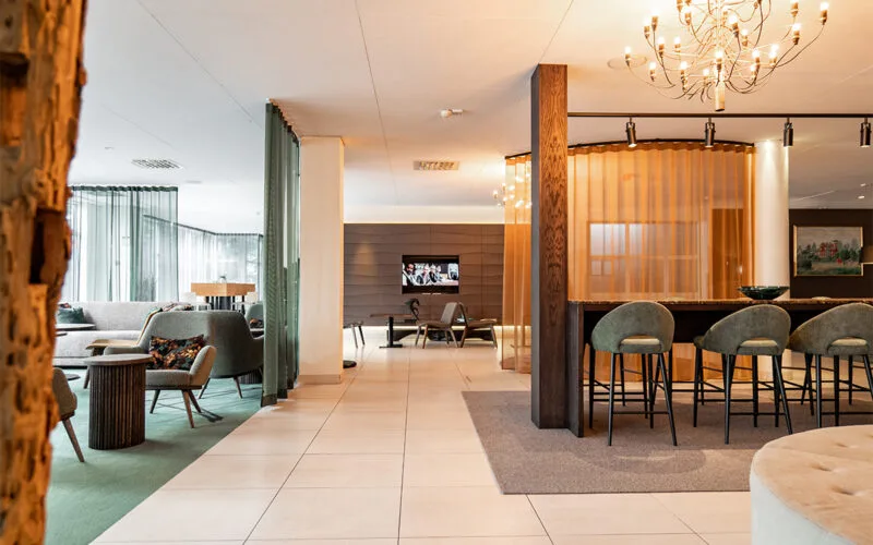 Hotellobby med moderna sittgrupper, högt bord med barstolar, mjuka gardiner och varm belysning.