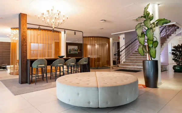 Hotellobby med stor rund sittmöbel, barstolar vid högbord, kristallkrona och trappa med mässingsräcken.