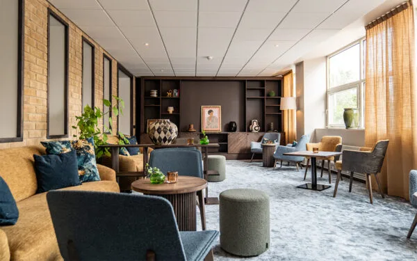 Hotellets lounge med sofaer, lænestole, runde borde og dekorative detaljer som planter og malerier, oplyst af store vinduer.