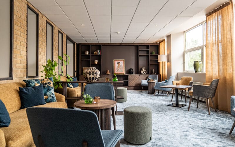 Hotellounge med soffor, fåtöljer, runda bord och dekorativa detaljer som växter och tavlor, belyst av stora fönster.