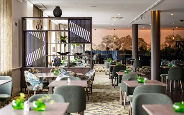 Restaurang med gröna stolar, mönstrade fåtöljer och träbord, avskärmat med dekorativ hylla.