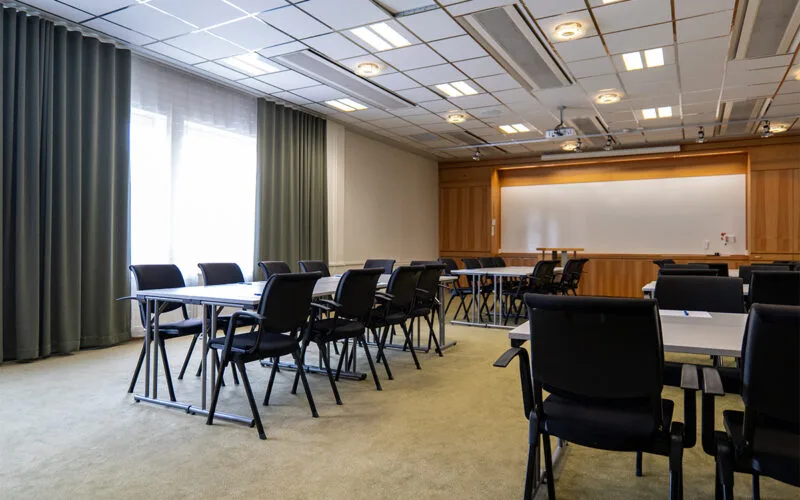 Konferensrum med uppställda bord och stolar, stora fönster med gardiner och whiteboard längst fram.
