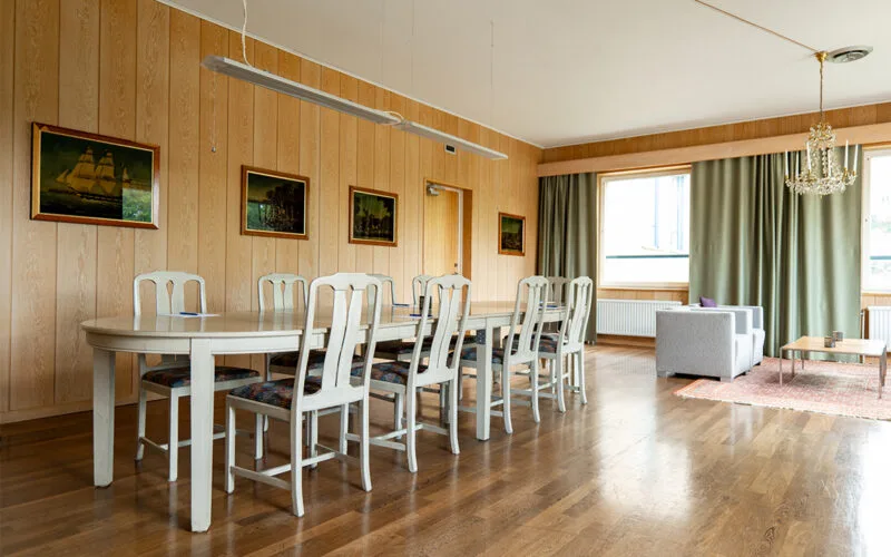 Mötesrum med konferensbord, vita stolar, tavlor på väggarna och loungehörna vid fönstren.