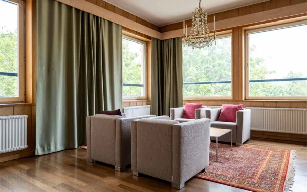 Loungehörna med fyra fåtöljer, soffbord, stora fönster, gröna gardiner och kristallkrona.