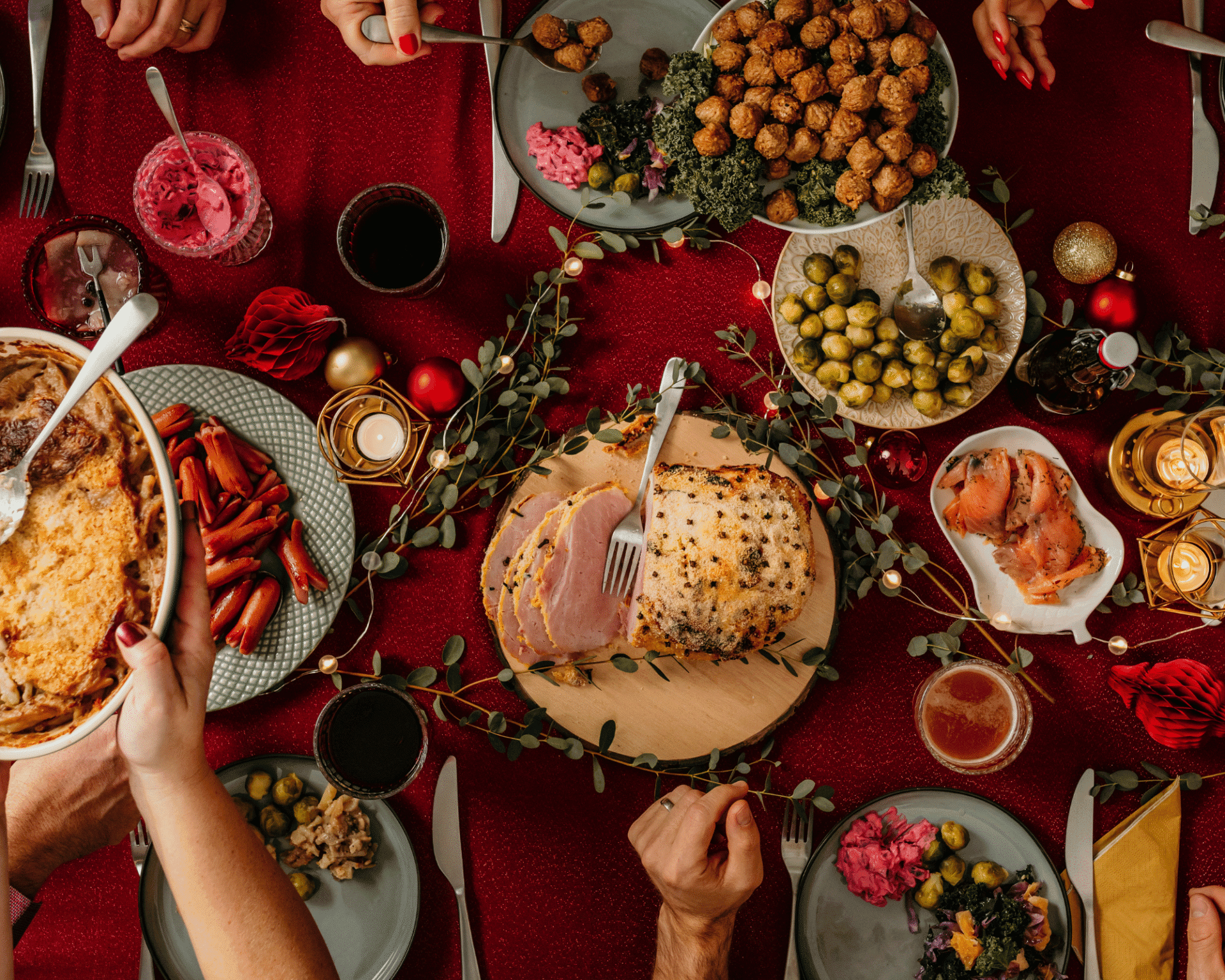 Traditionellt julbord med julskinka, köttbullar, prinskorv, Janssons frestelse, lax och olika tillbehör på ett rött dukat bord med juldekorationer.