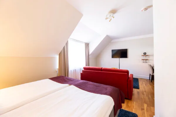 Hotellrum med dubbelsäng, röd soffa och TV placerad på väggen