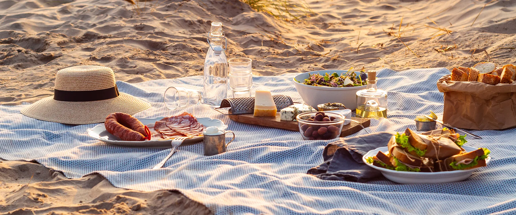 Picknick på stranden med filt, mat, dryck och stråhatt.