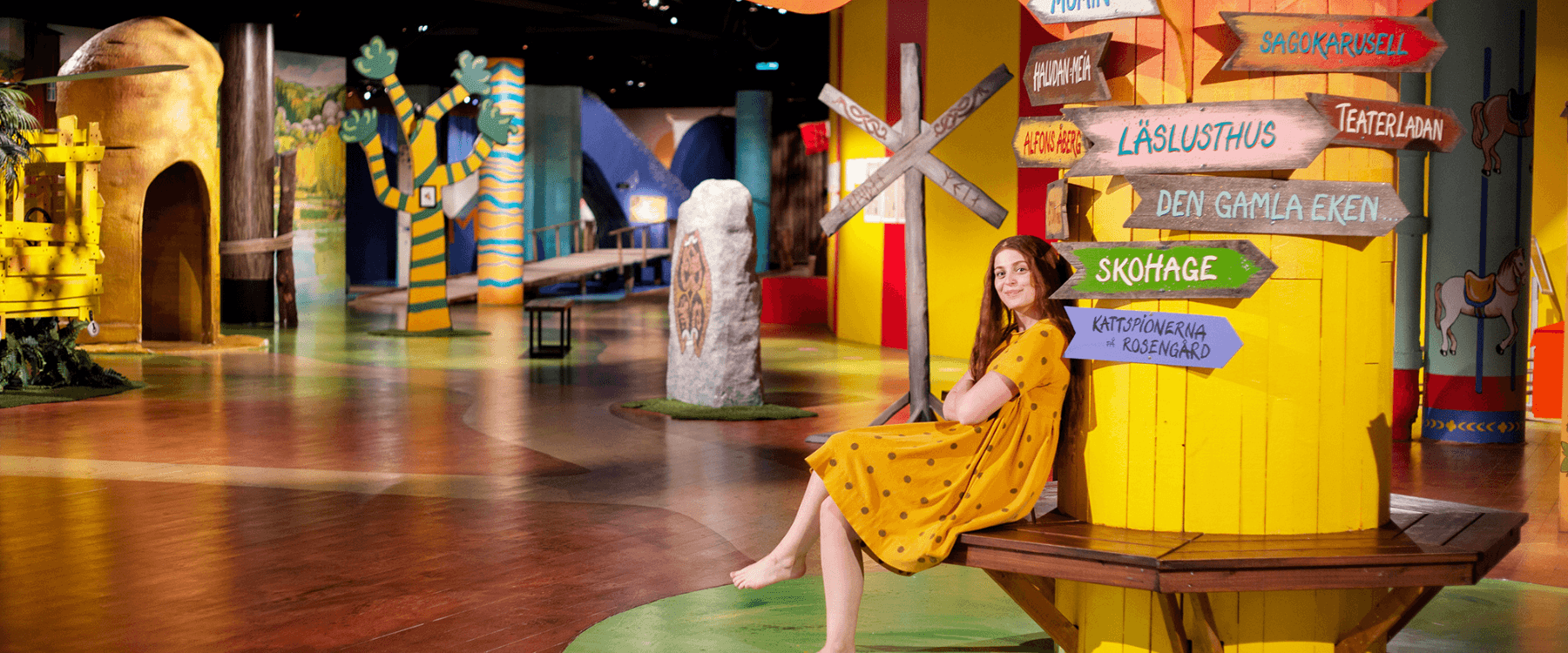 En kvinna i gul klänning sitter barfota på en bänk runt en färgglad, målad trädstam inomhus. Trädet har skyltar som pekar mot olika sagolika platser, som ”Muminhuset” och ”Sagokarusell”. Runt omkring syns färgglada dekorationer och kulisser i en fantasifull miljö.