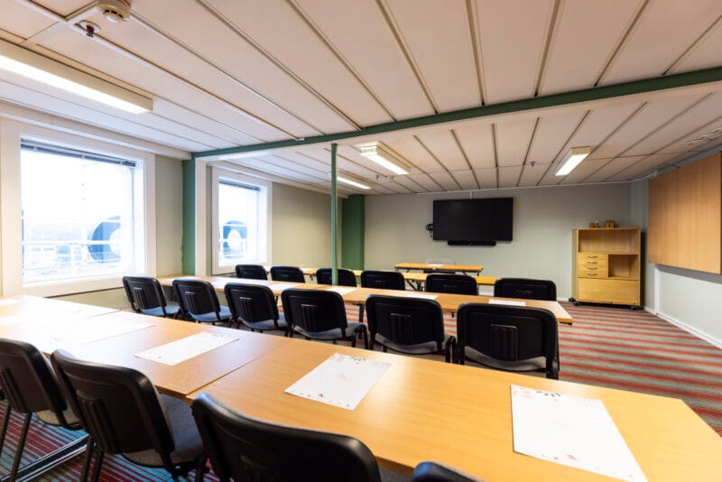 Konferensrum med mötesbord och grå stolar, dukat med glas och block, samt whiteboard och projektor i framkant.